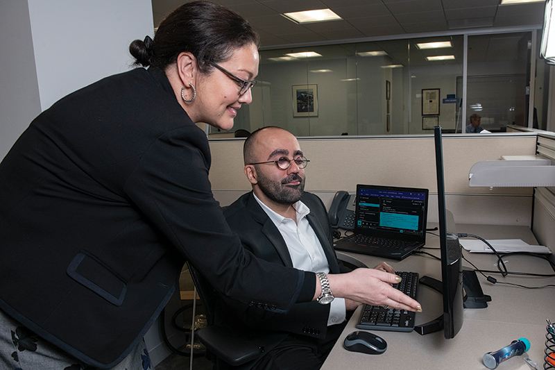 A White woman smiles as she looks at a screen with a Middle Eastern man with glasses who is seated at his desk. 