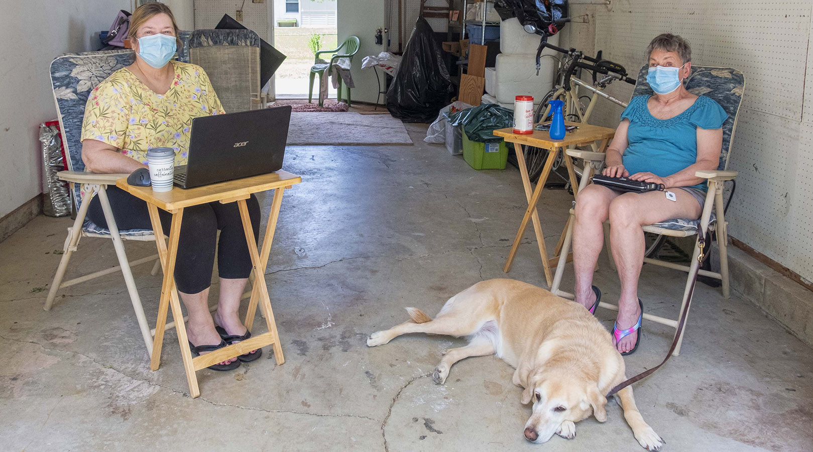Two White women are socially distanced in a garage, both wearing masks -- one woman uses a braille notetaker as a dog lies at her feet and the other uses a laptop.