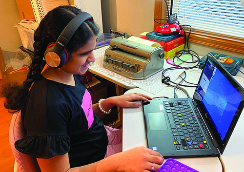 An Indian girl laughs as she works at a computer in her home. The keyboard has colorful tactile stickers on it. A Perkins brailler is on the desk to her left, positioned under a document camera. She is wearing headphones.