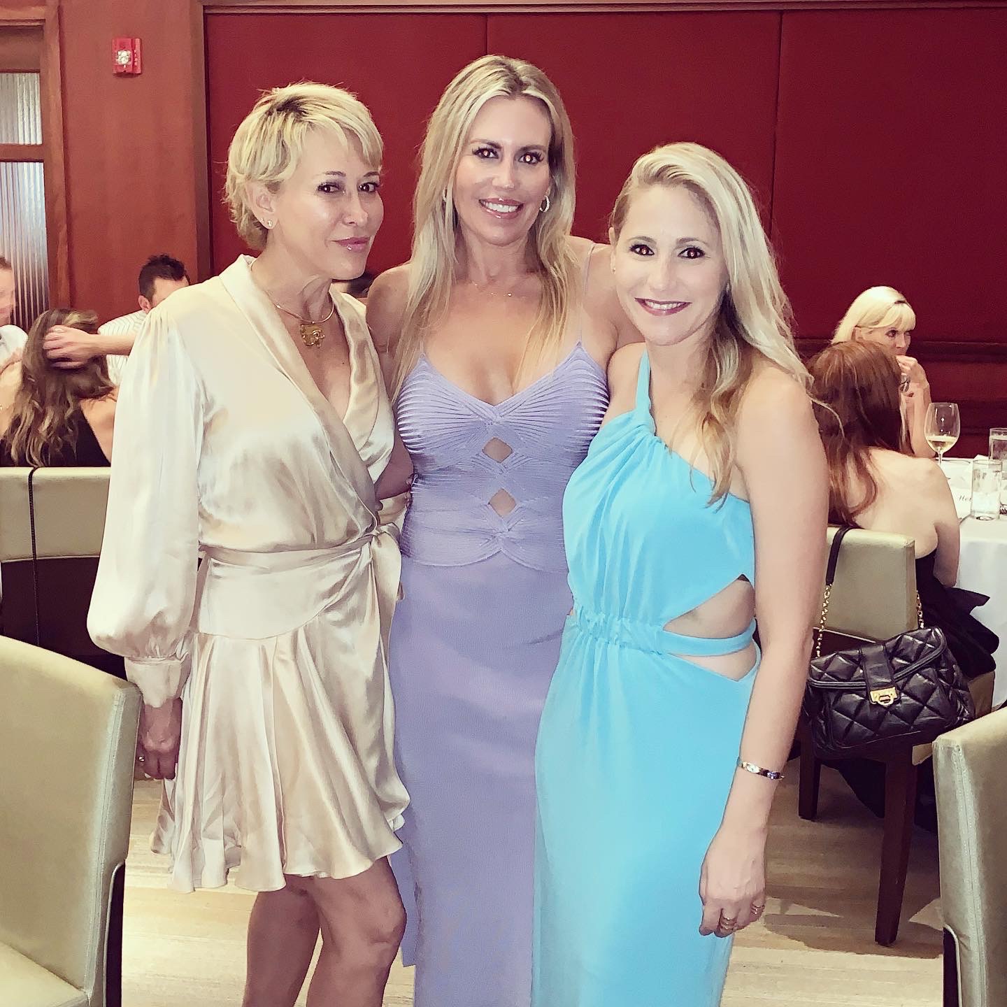 A group of three white blonde women in fancy dresses smile for the camera, standing in front of tables full of people.