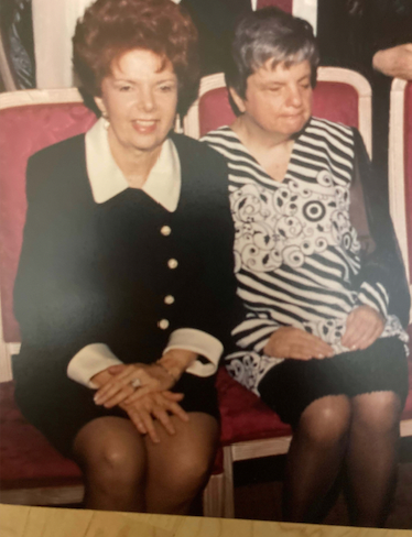 the image shows Marilyn and Francine Gruder sitting next to each other. Marilyn sits on the left with red hair and is wearing a black dress. Next to her is Francine, who is wearing a dress with stripes. Both sisters are smiling. 