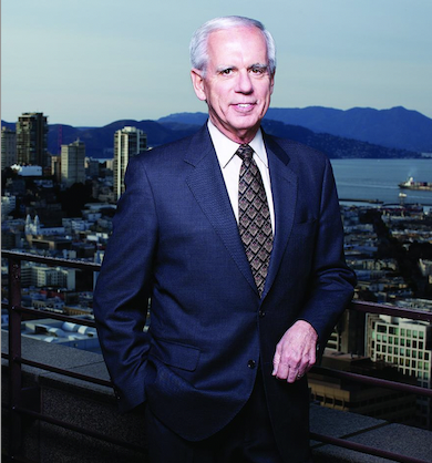 Tony Coelho is wearing a blue suit and is standing behind an image of a city. He is smiling. 