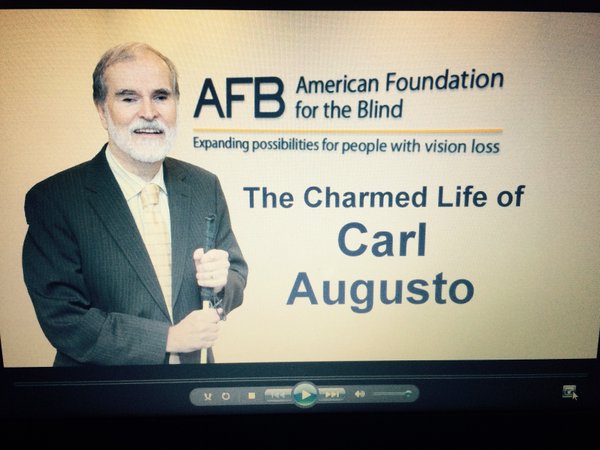 still image from the video honoring The Charmed Life of Carl Augusto - showing Carl, cane in hand, in front of the American Foundation for the Blind logo