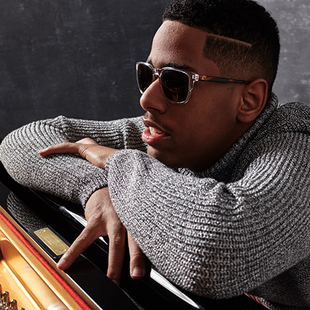 Matthew Whitaker, a young black man wearing sunglasses and a gray sweater. His crossed arms are resting on a piano keyboard.