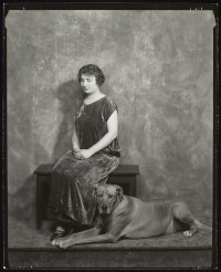 Photograph of Helen Keller seated on a bench indoors with a large dog seated beside her.; circa 1925