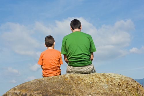 A father and son sit on a rock outcrop overlooking the sky.