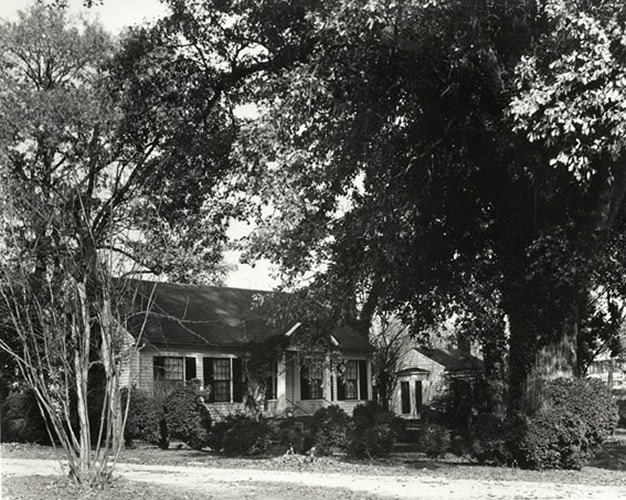 Photo of a white house, one-story at the front with a small porch, surrounded by trees, with a smaller house at the side