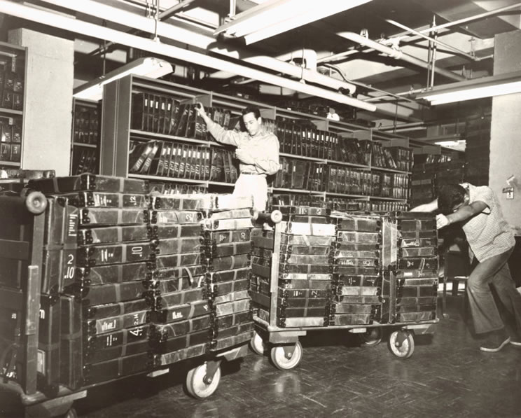 This photograph was taken in the shipping department at the Library for the Blind, New York Public Library, and shows Talking Book records being mailed in their containers. In the foreground of the photograph a man pushes two large platforms on wheels full of Talking Book records in their containers. In the background a man is standing on a ladder in front of one of the many large metal shelves housing the containers. Talking Book Archives, American Foundation for the Blind.