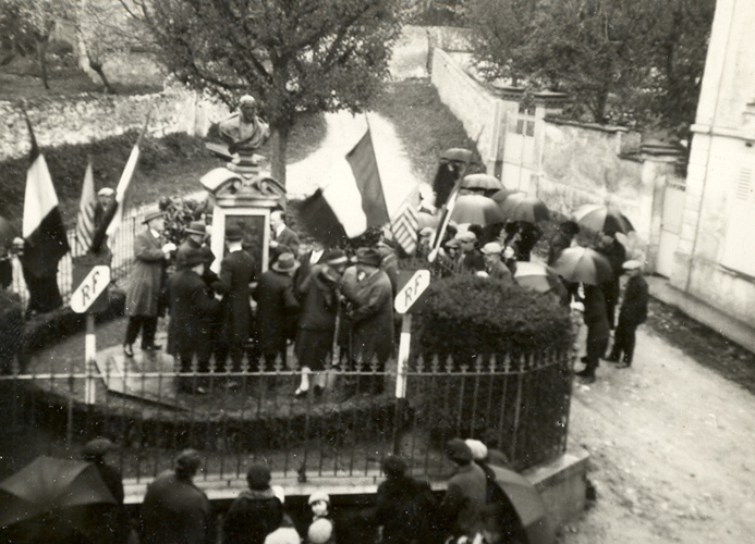 Photograph of a crowd standing around Louis Braille's monument in Coupvray in 1929. The photographer is above the scene looking down and along the village street. The monument is a bust of Louis. The bust rests on a pedestal within a small fenced off area in the middle of the street. French flags and signs with the letters RF for Republique Francaise are attached to posts. Citizens carry umbrellas.