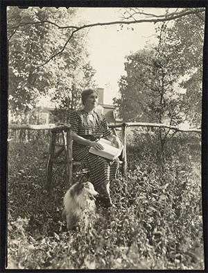 Helen Keller sitting and reading braille in the garden with her collie dog standing next to her.