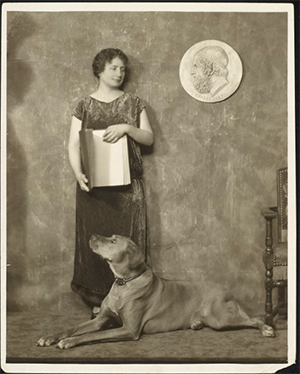 Circa 1925, Helen Keller stands, possibly in the photographer's studio, with a dog (possibly Sieglinde) seated on the ground in front of her. She is seen looking slightly downwards and is holding a book which is open to the viewer. She is wearing a shimmery, patterned, full-length dress that is loose and has small cap-sleeves and a boat neck. The dog is looking up at Helen Keller. A circular bas-relief with a profile of Homer is on the wall to Keller's left, a chair is also just visible in the image.