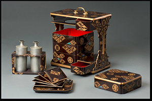 a red and black lacquer New Year's luncheon set that was given to Helen as a gift from Kazuo Honma, founder of the Japanese Braille Library. The main box is in black lacquer with gold symbols and carved abalone inlays, all set in carrying stand with brass top handle. Six drawers (boxes), one with cover, one medium size tray, five small ones, removable flask holder, pair of pewter cylinder flasks with top handles inlaid with red glass. Interior of drawers in red lacquer.