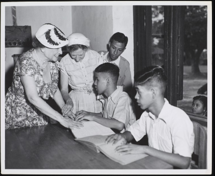 Photograph taken indoors of Helen Keller and Polly Thomson visiting students reading braille at the St. Michael's School for the Blind, Rangoon, Burma (Myanmar). 5/15/1955
