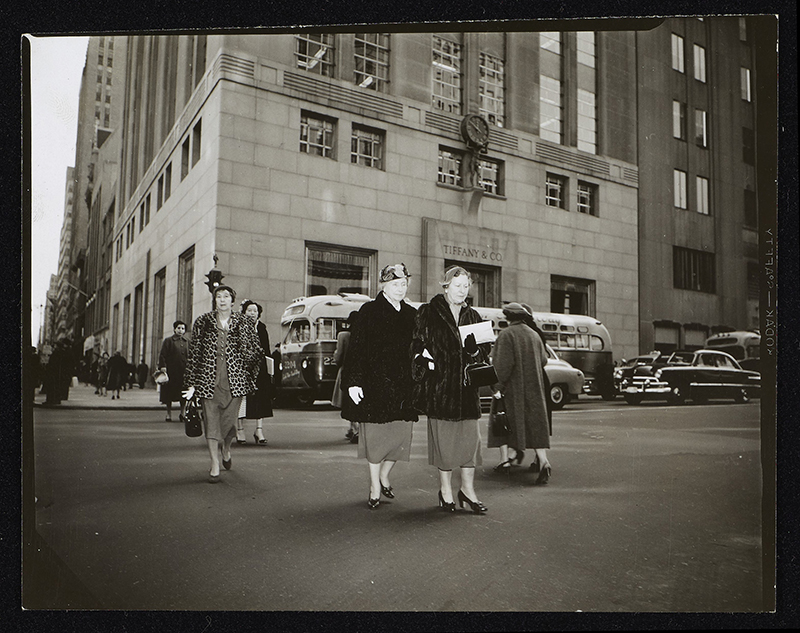 Helen Keller and Polly Thomson walk arm in arm across Fifth Avenue at 57th Street in New York City. The store Tiffany & Co. is visible in the background, as are a bus and cars from that period. Helen and Polly both wear dark three-quarter length fur coats, below-the-knee length skirts and hats.