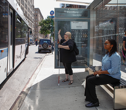 Two women waiting at a bus stop. One woman is sitting. The other is standing and hailing the public transit bus is it pulls into the stop. 