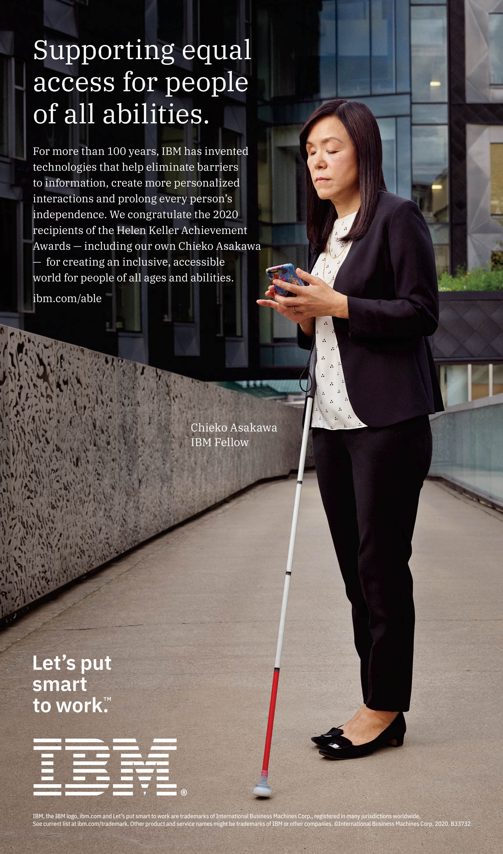 Image: Chieko Asakawa, IBM Fellow, stands in the center of an outdoor walkway, surrounded by office buildings. Her white cane is propped against her body as she uses her smartphone.