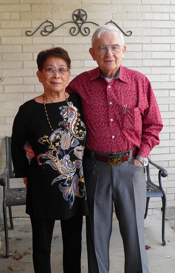 Matt and his wife Kay, standing outside in front of a brick wall. Matt has his arm around Kay, and both are smiling at the camera.