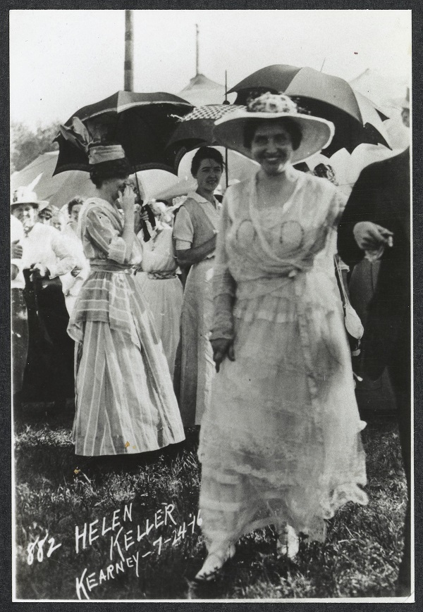 Taken outdoors. Helen Keller with a group of women standing behind her holding parasols.