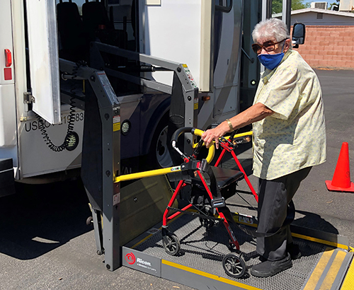 An older White woman wearing a mask and using a walker stands on the boarding lift to a paratransit van.