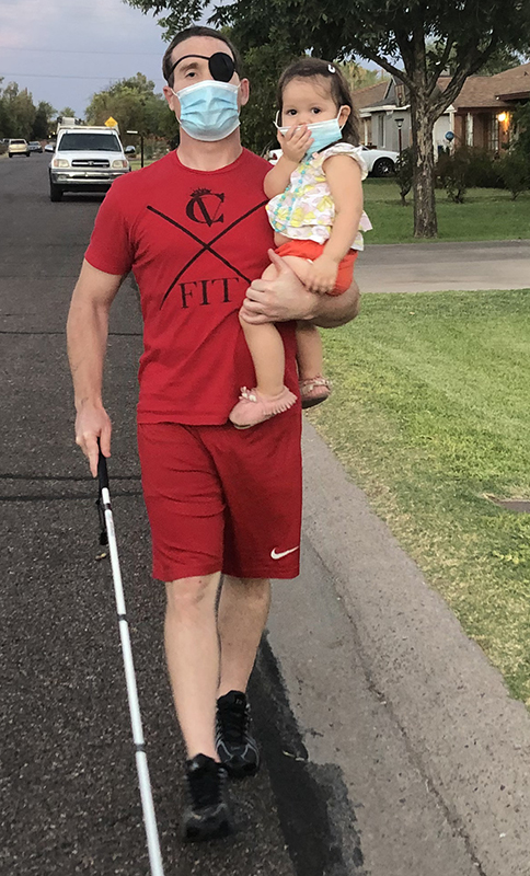 A White man wearing a mask and carrying a baby uses a white cane as he walks down a residential street.