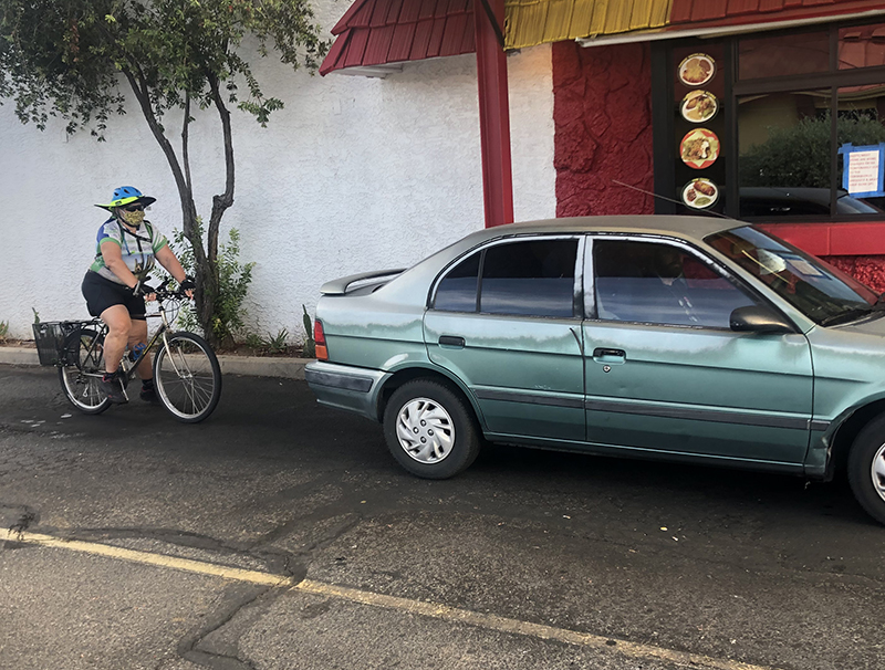 A White woman wearing a mask and riding a bicycle waits in a restaurant’s drive-through line behind a car. 