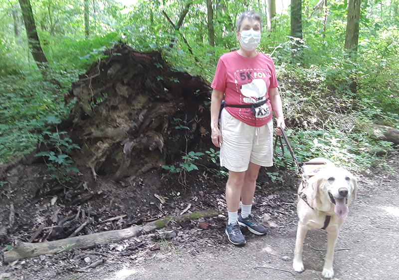 A middle-aged White woman wearing a mask stands in a park with her dog guide.