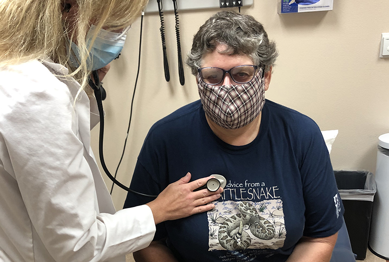 In an examination room, a White female doctor wearing a mask uses a stethoscope to listen to the heart of an older White woman.