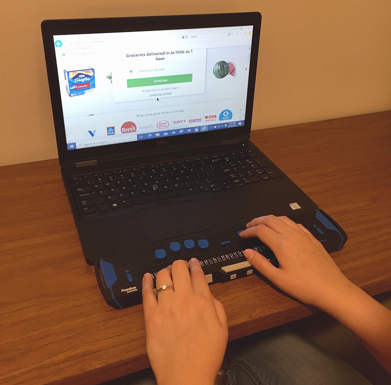 A White woman uses a laptop with a refreshable braille display to order groceries.