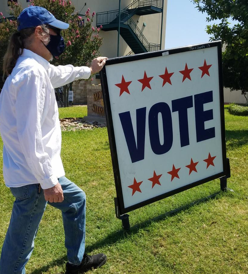 A middle-aged White man wearing a mask stands next to a sign that reads “VOTE.”