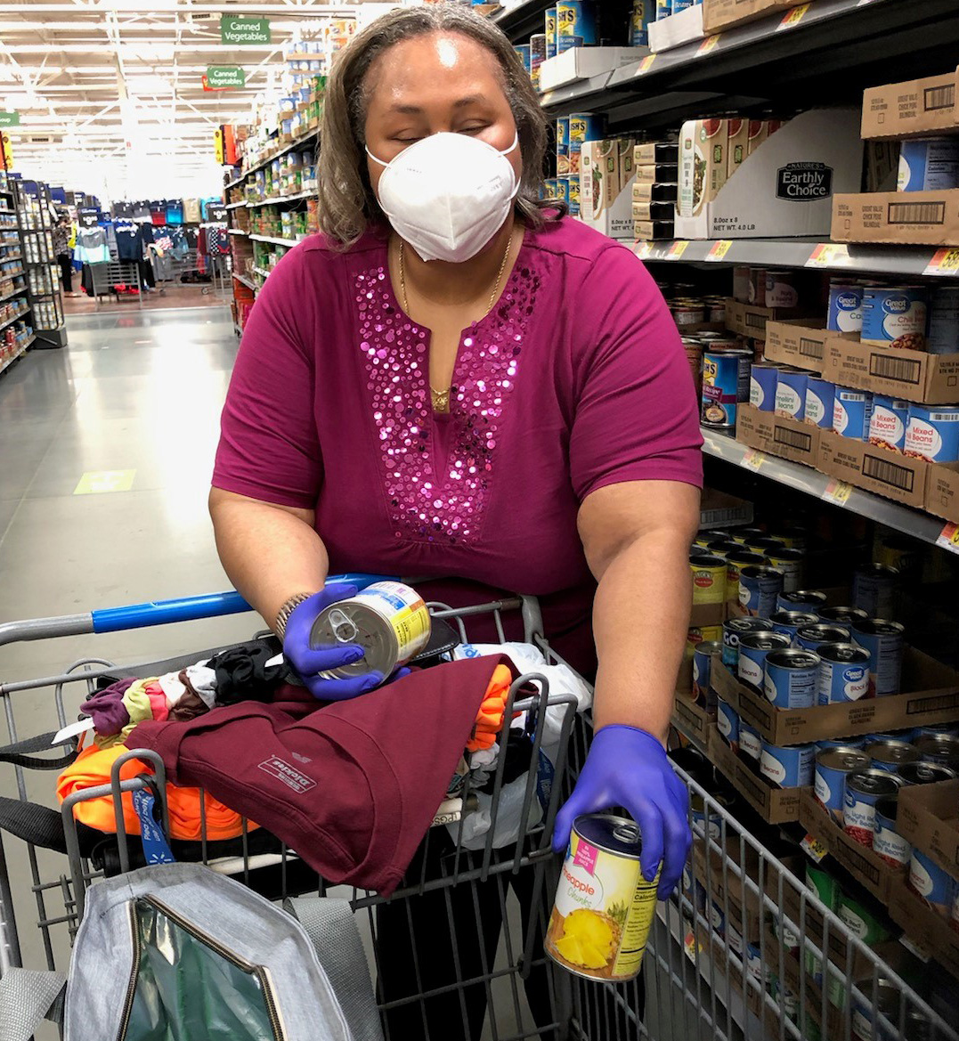 A middle-aged Black woman wearing a mask and nitrile gloves shops for groceries. She is placing a can in a shopping cart. 