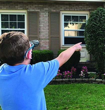 A school-age White boy wearing a mask uses a moncular in his left hand and with his right hand points to a house number.