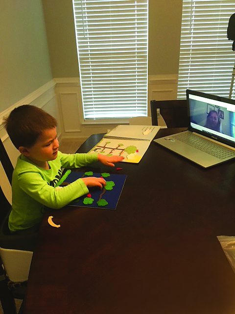 A white preschool boy sits at a table. With his right hand he explores a felt tree that accompanies the book “Best for a Nest”. His left hand is on the tree in the book. A woman is visible on a laptop watching him.