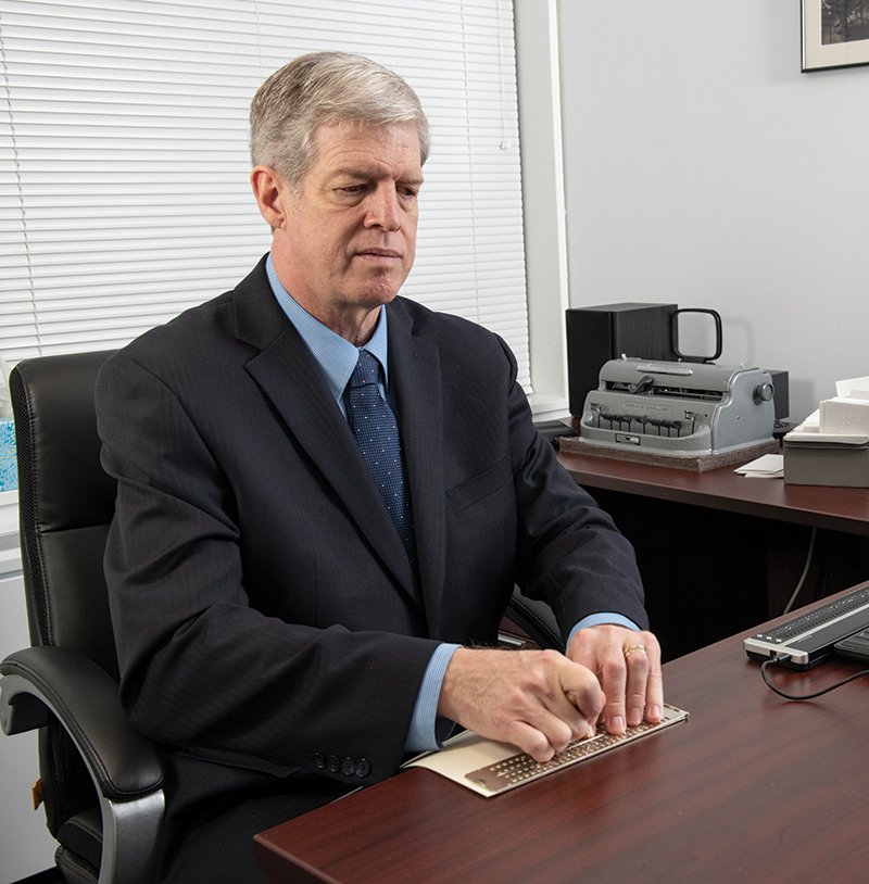 Kirk Adams sits at his desk with a braille display