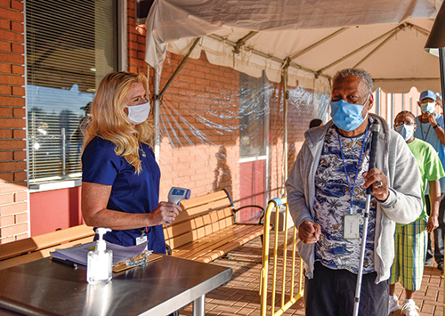 A White woman wearing a mask holds a thermometer and addresses a Black man wearing a mask and holding a white cane. He stands at the front of a line of people in a covered outdoor area. 