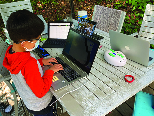 A young boy wearing a mask and wearing glasses uses a laptop at an outdoor table covered with laptops, tablets, and a braille display. 