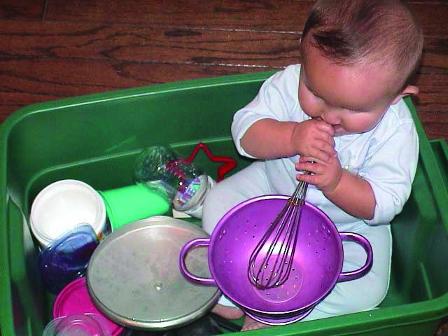 A White infant boy with additional disabilities sits in a plastic tub full of kitchen supplies. He has the end of a whisk in his mouth as he places it in a metal colander.
