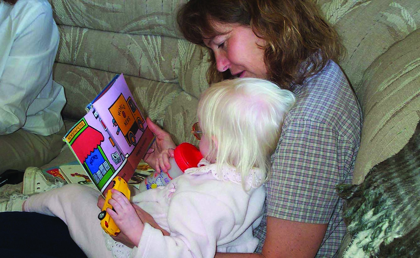 A White mother and her toddler girl with albinism explore a book together about vehicles. The toddler holds a yellow plastic car in her left hand. 