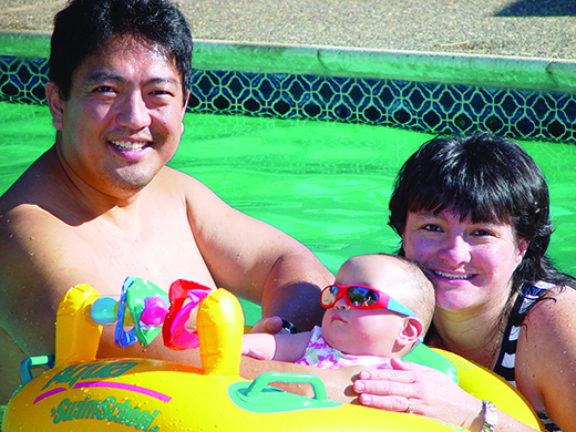 An Asian family are in a swimming pool. The infant daughter wearing sunglasses is placed in a yellow floatie. The mother and father are smiling broadly. 