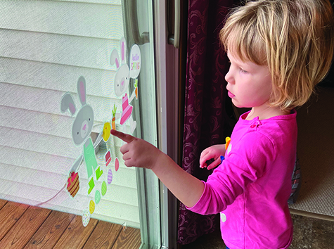 A White preschool girl stands at a sliding glass door decorated with Easter decals. She points to a carrot. 