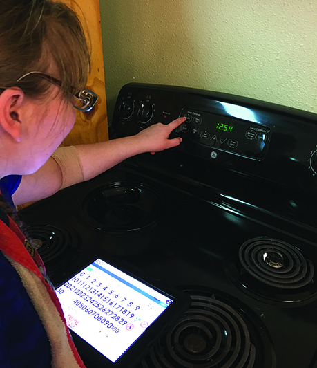 A White school-age teenage girl with additional disabilities sets the temperature on an oven. Her iPad with large print rests on the stove. 