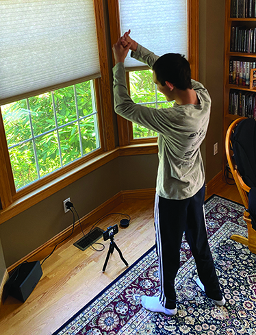 In a livingroom, a White teenage boy with additional disabilites wearing workout clothes stands in front of a smartphone on a stand with a large speaker sitting next to it on the floor. His arms are extended over his head as he exercises. 