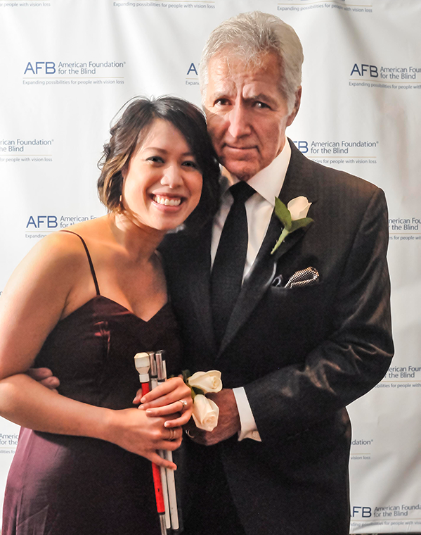 Alex Trebek stands next to Christine Ha. His right arm is around her waist and his left hand holds hers in front of them. Christine has her white can folded up and in her hands. Alex is wearing a grey suit and tie. Christine is wearing a burgundy dress with spaghetti straps.