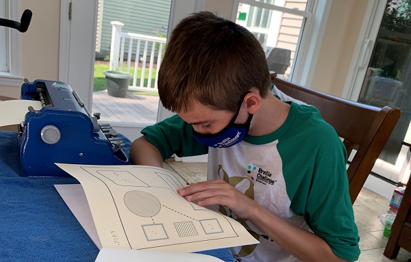 Boy sits at table next to braille embosser. He is looking through tactile graphs. He wears a mask to avoid spreading viruses.