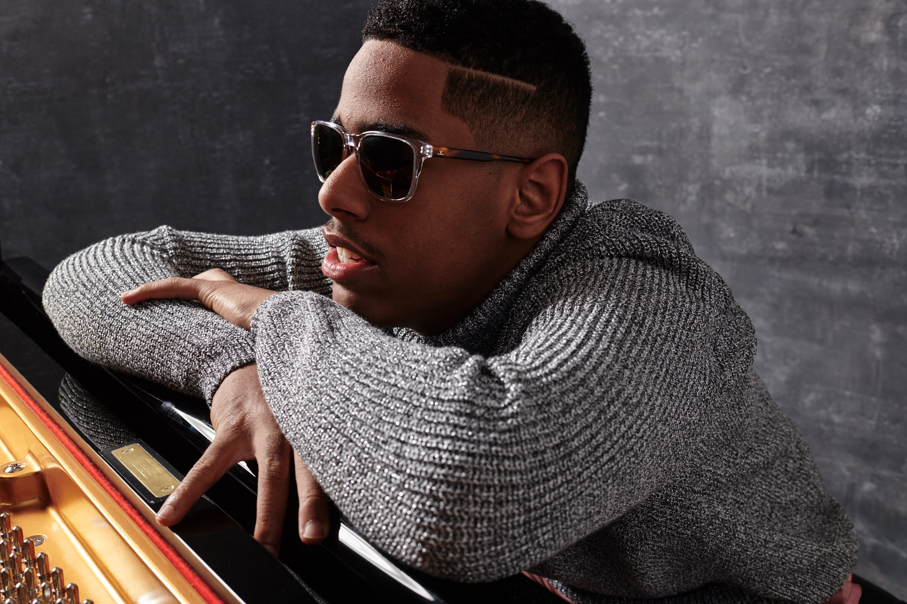Matthew Whitaker, a young black man wearing sunglasses and a gray sweater, leans on a piano keyboard