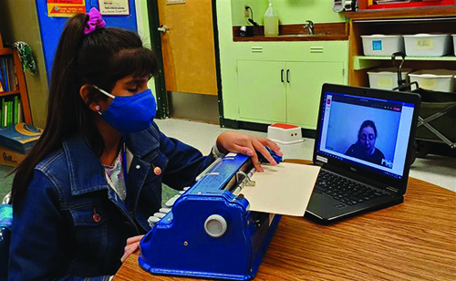 A Hispanic girl wearing a mask reads what she wrote on a Perkins braille writer. An instructor looks on through the computer screen. The girl is in a classroom. 