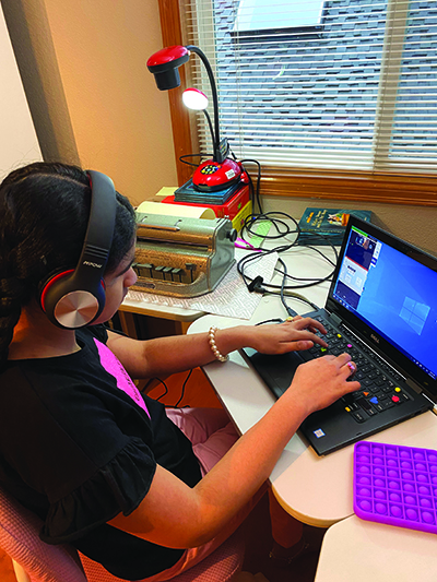 An Indian girl laughs as she works at a computer in her home. The keyboard has colorful tactile stickers on it. A Perkins brailler is on the desk to her left, positioned under a document camera. She is wearing headphones. 