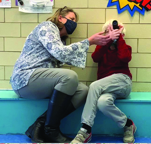 A White, elementary school-aged boy with albinism looks through a monocular with his White female TVI supporting his hand position on the monocular. They are both wearing masks and sitting on a bench in a school hallway. 