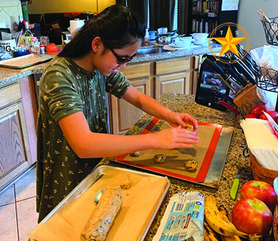 A Chinese American girl places cookie balls on a cookie sheet as her teacher and classmate look on via Zoom from a device to her left. On her right she has more cookie dough. 
