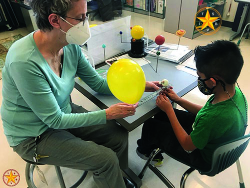 In a classroom both a Hispanic elementary student and a White teacher wear masks as they measure the distance between a big yellow balloon and a small ball about 10 inches away. The balloon represents the sun and the ball a planet. 