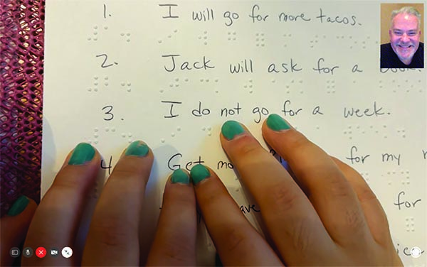 A Latina girl whose nails are painted green reads brailled sentences as her White male TVI smiles as he watches via Zoom. 